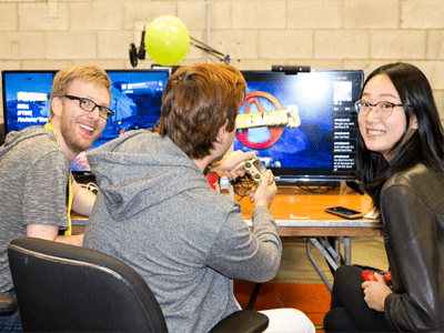 Three people sitting in front of a video game monitor, turned to face the camera smiling.