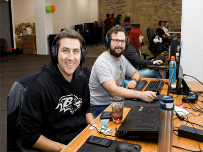 Two participants sitting at desks playing video games while smiling at camera.