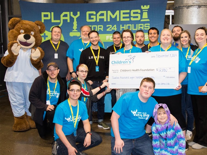 Group of people wearing Extra Life shirts crowded around a large cheque. There is a bear mascot in blue scrubs beside them waving.